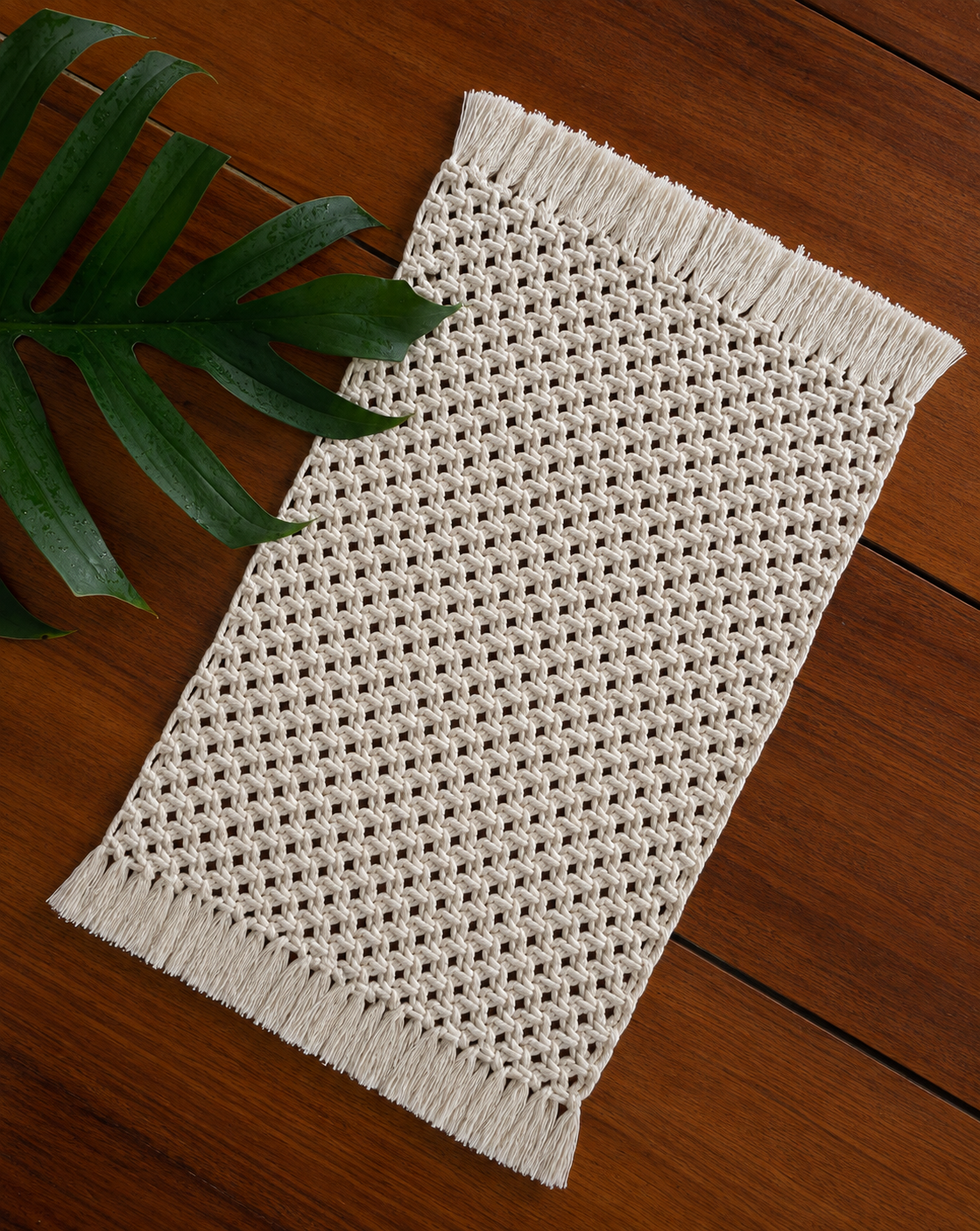 Jogo americano retangular, em tom cru, confeccionado em macramê com fios trançados em uma trama vazada e delicada. A peça possui franjas longas e uniformes nas duas extremidades menores, combinando textura artesanal e aparência natural.